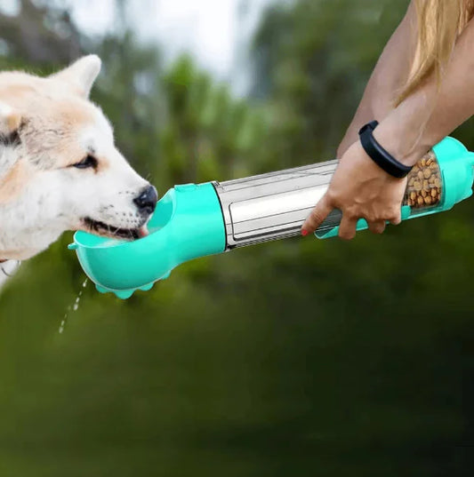 Dog drinking water from a green and silver portable water fountain held by a person outdoors.