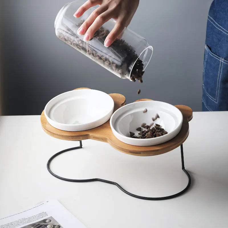 Person pouring food into a modern pet feeder with two bowls on a white surface.