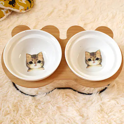 Two white bowls with cat illustrations on a wooden stand on a fluffy surface.